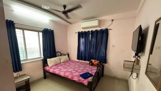 Quarto com cama, ventoinha no teto, ar condicionado, janela com cortinas, mesa pequena e televisão.