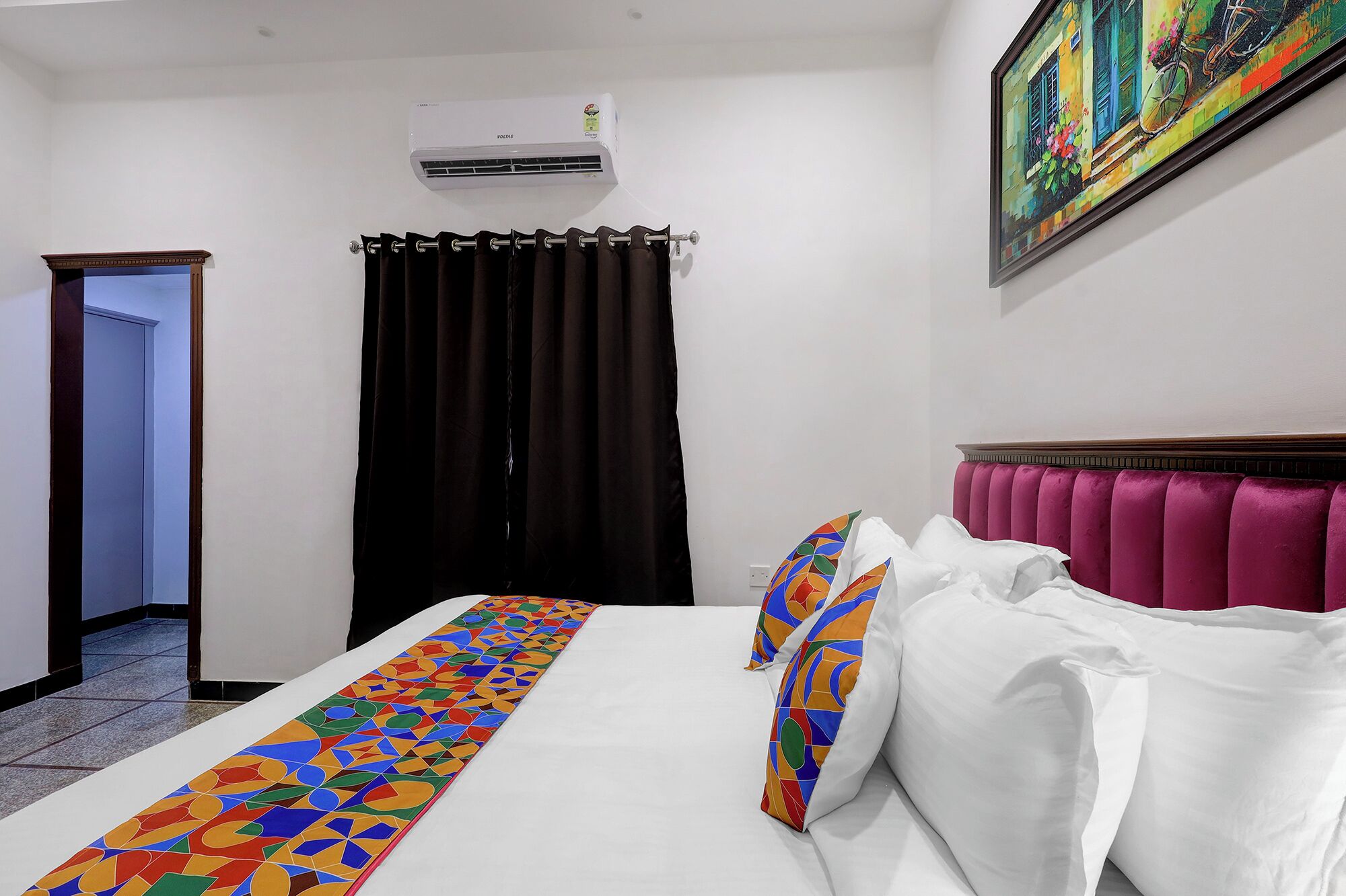 A hotel room with a bed, a window with a curtain, an air conditioning unit, and a painting on the wall.