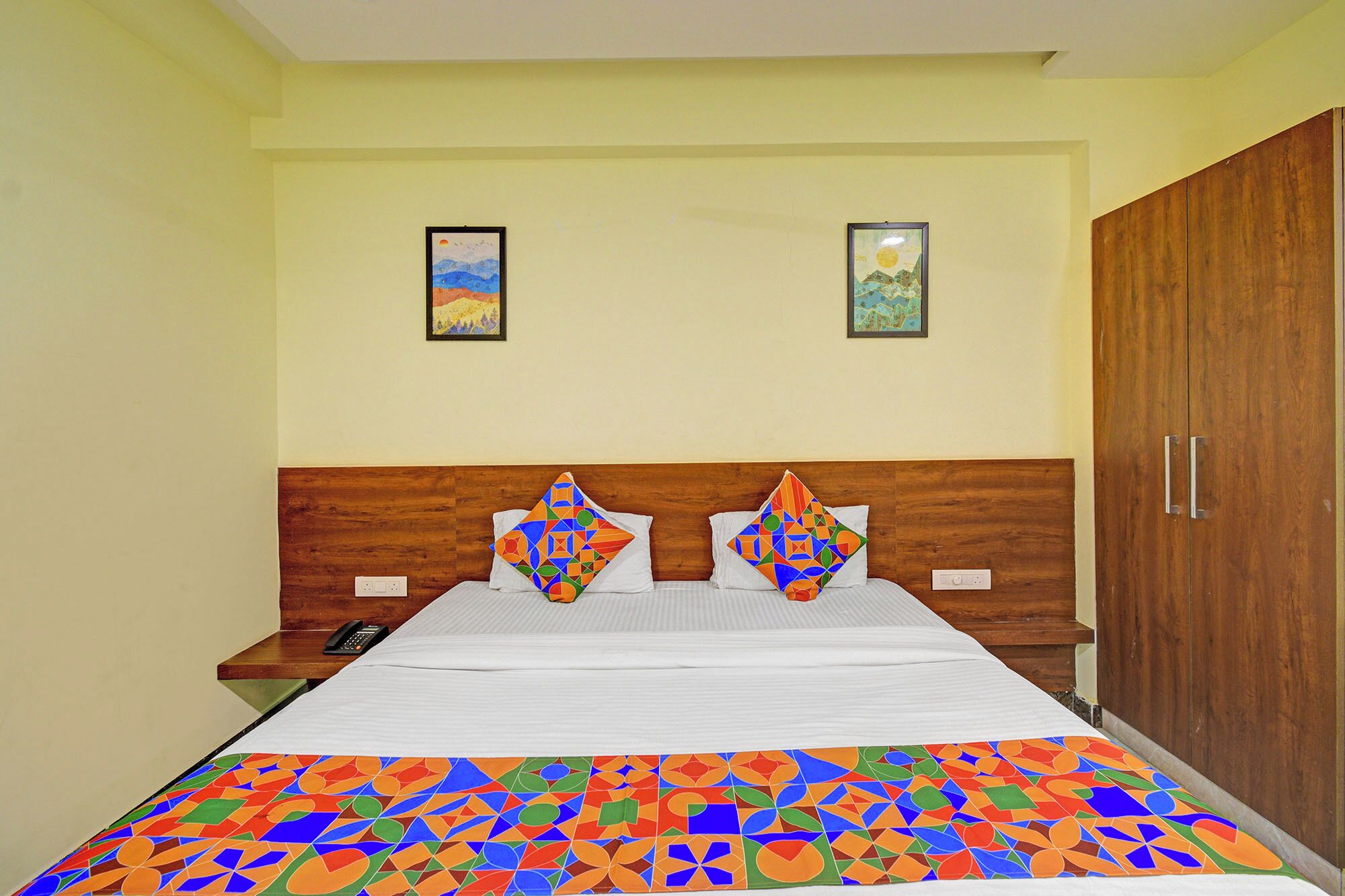 A hotel room with a wooden headboard, a bed with a colorful geometric-patterned bedspread, two framed pictures on the wall, and a wooden wardrobe.
