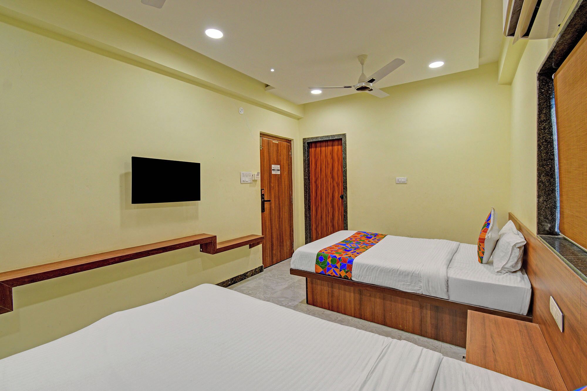 A hotel room with two beds, a ceiling fan, a flat-screen TV mounted on the wall, and a wooden door.