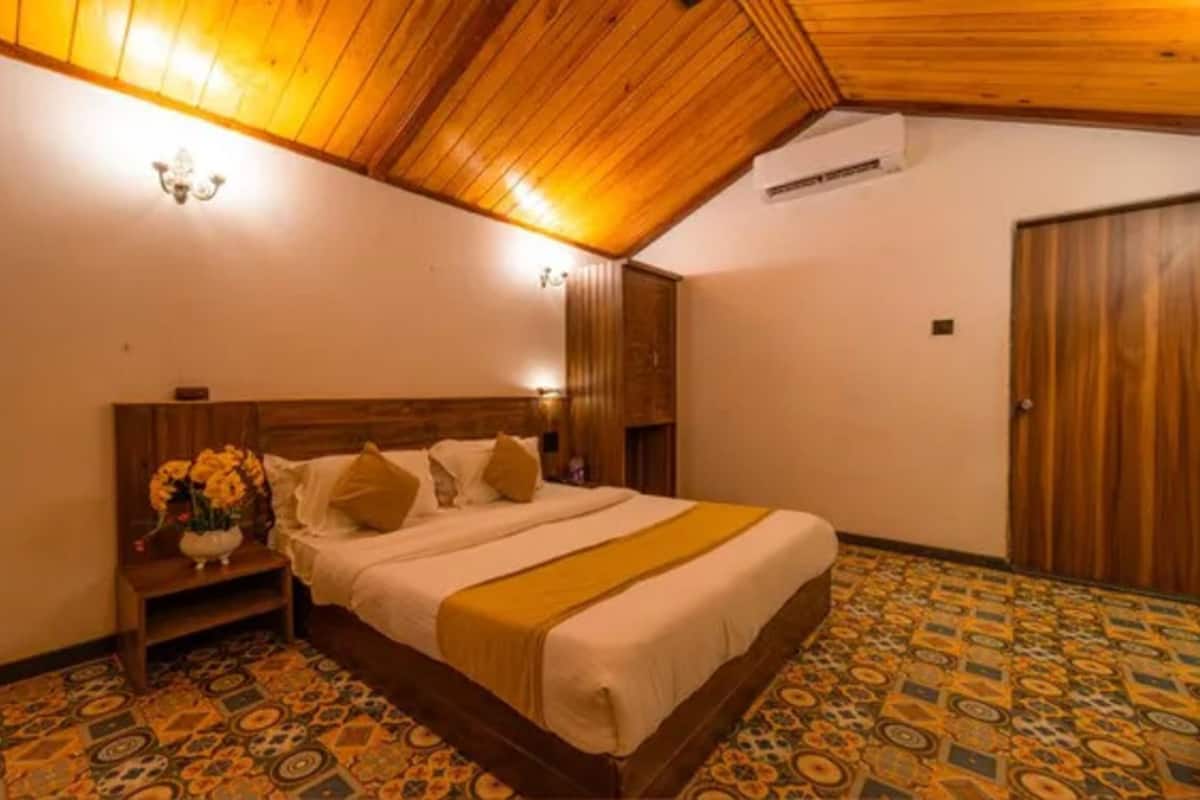 A bedroom with a wooden ceiling, a patterned floor, a bed with a yellow throw, and a nightstand with a vase of flowers.