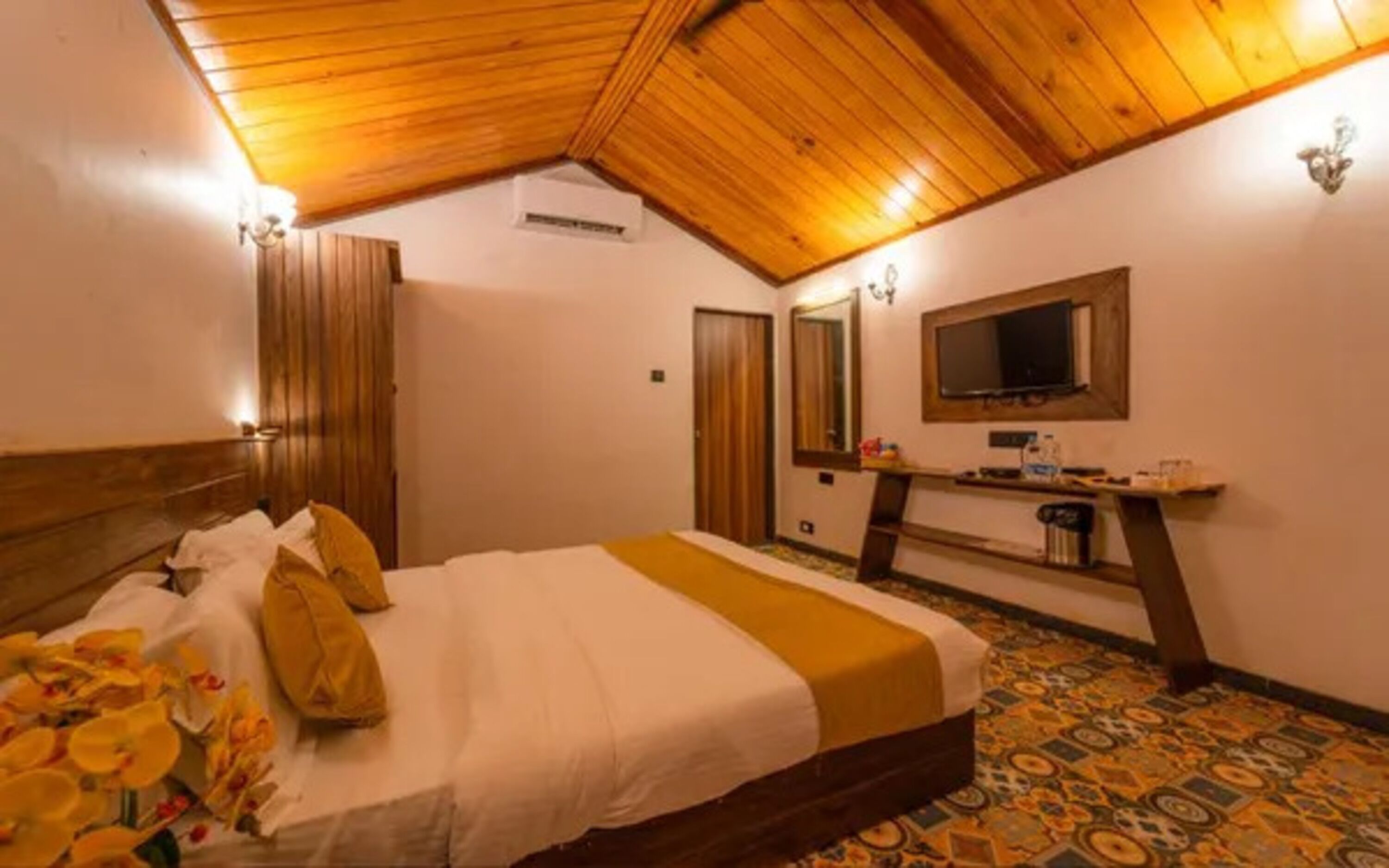 Ein Hotelzimmer mit Holzbalkendecke, einem Bett mit weiß-gelber Bettdecke, einem kleinen Schreibtisch mit Spiegel und einem Fernseher darüber.