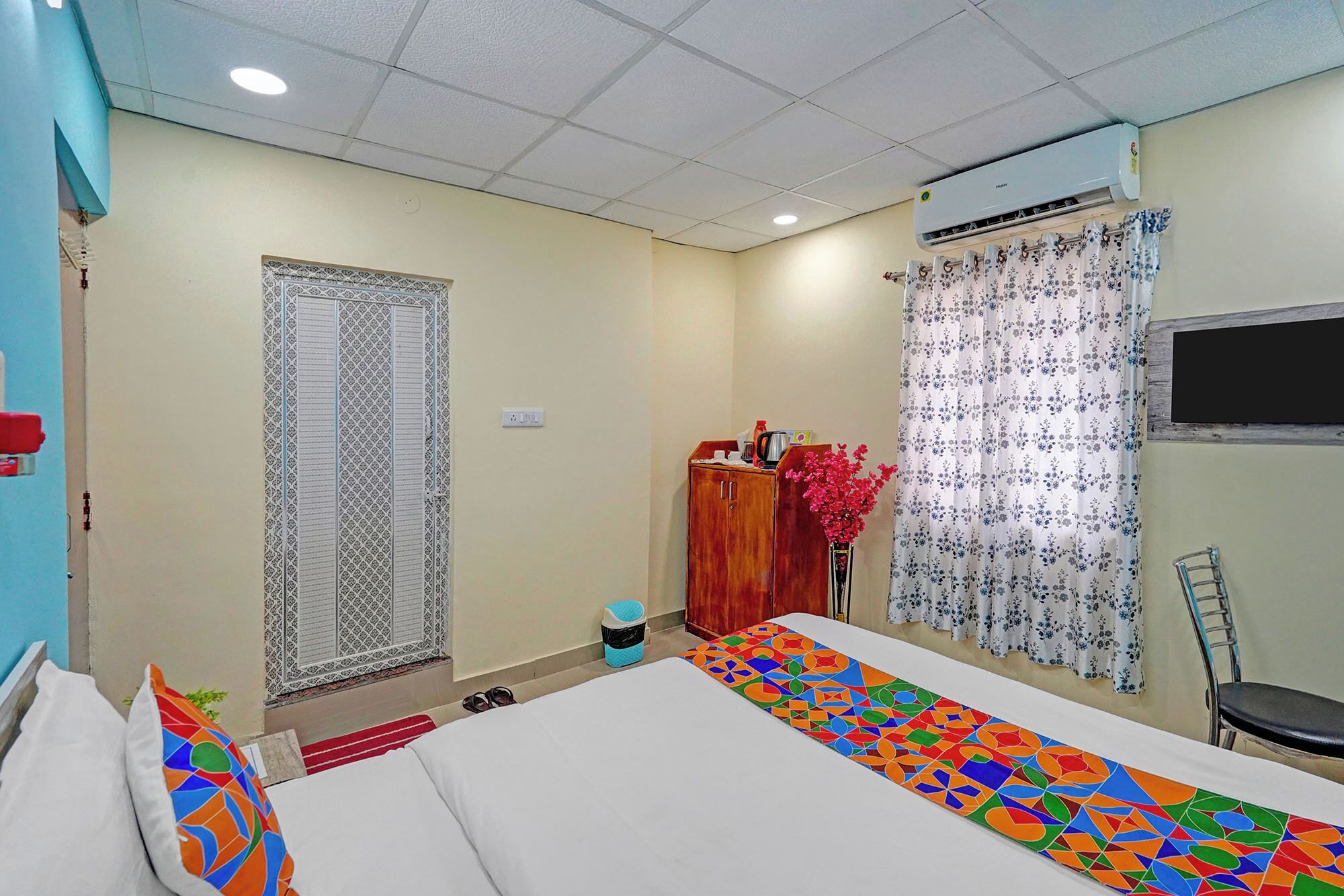 A hotel room with a bed, a desk, a chair, an air conditioner, a window with curtains, and a door with a patterned design.
