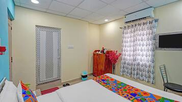 A hotel room with a bed, a desk, a chair, an air conditioner, a window with curtains, and a door with a patterned design.