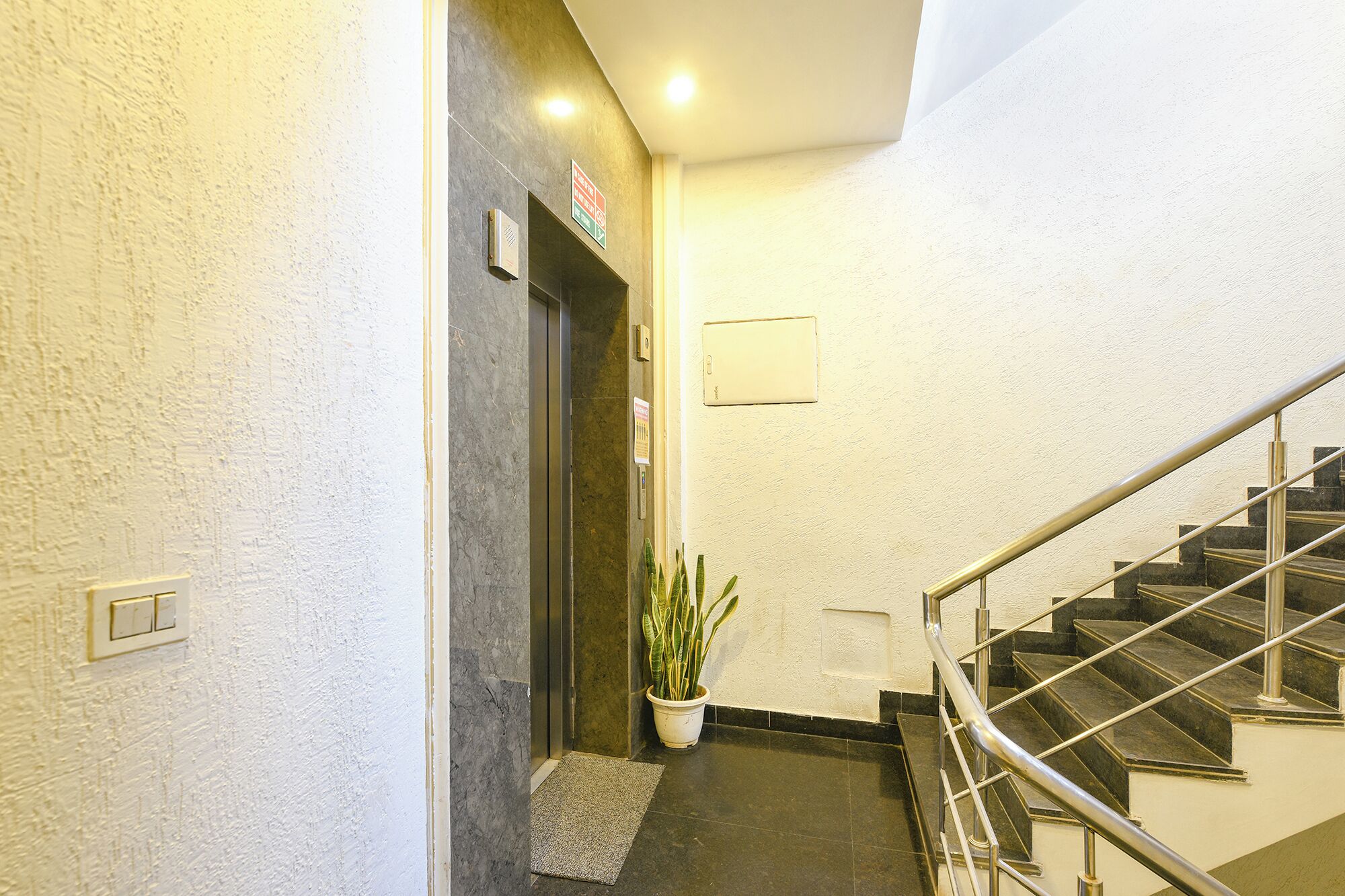 A hotel lobby with a staircase, a potted plant, a fire alarm, and a light switch on the wall.