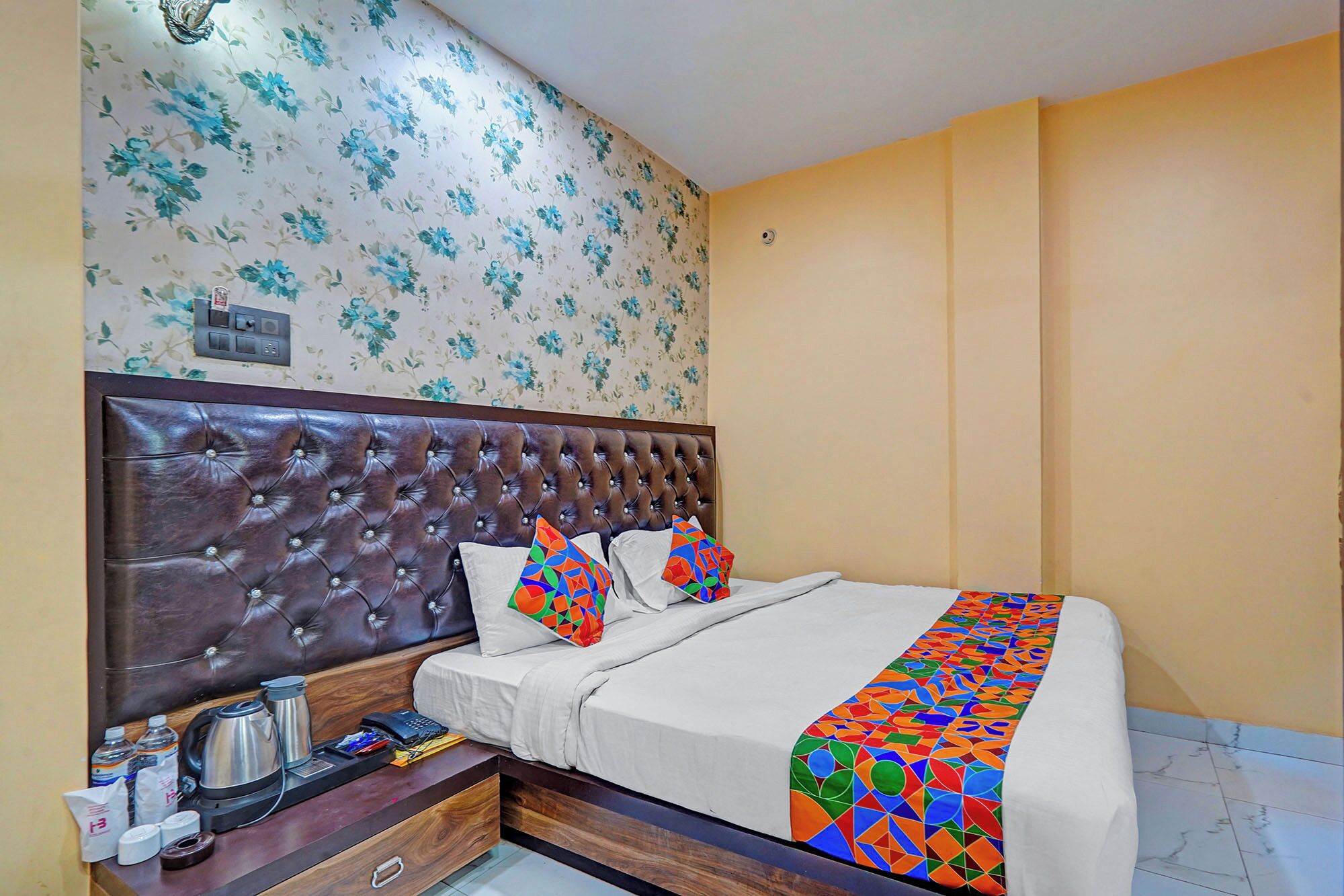 A hotel room with a tufted headboard, floral wallpaper, a bed with colorful pillows, and a bedside table with a kettle and a phone.