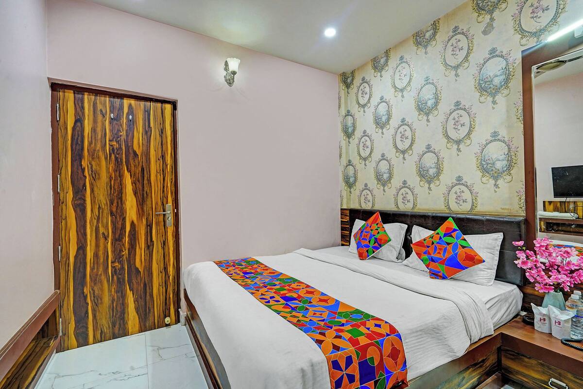 A bedroom with a wooden door, a bed with colorful pillows, a patterned wallpaper, and a nightstand with a vase of flowers.