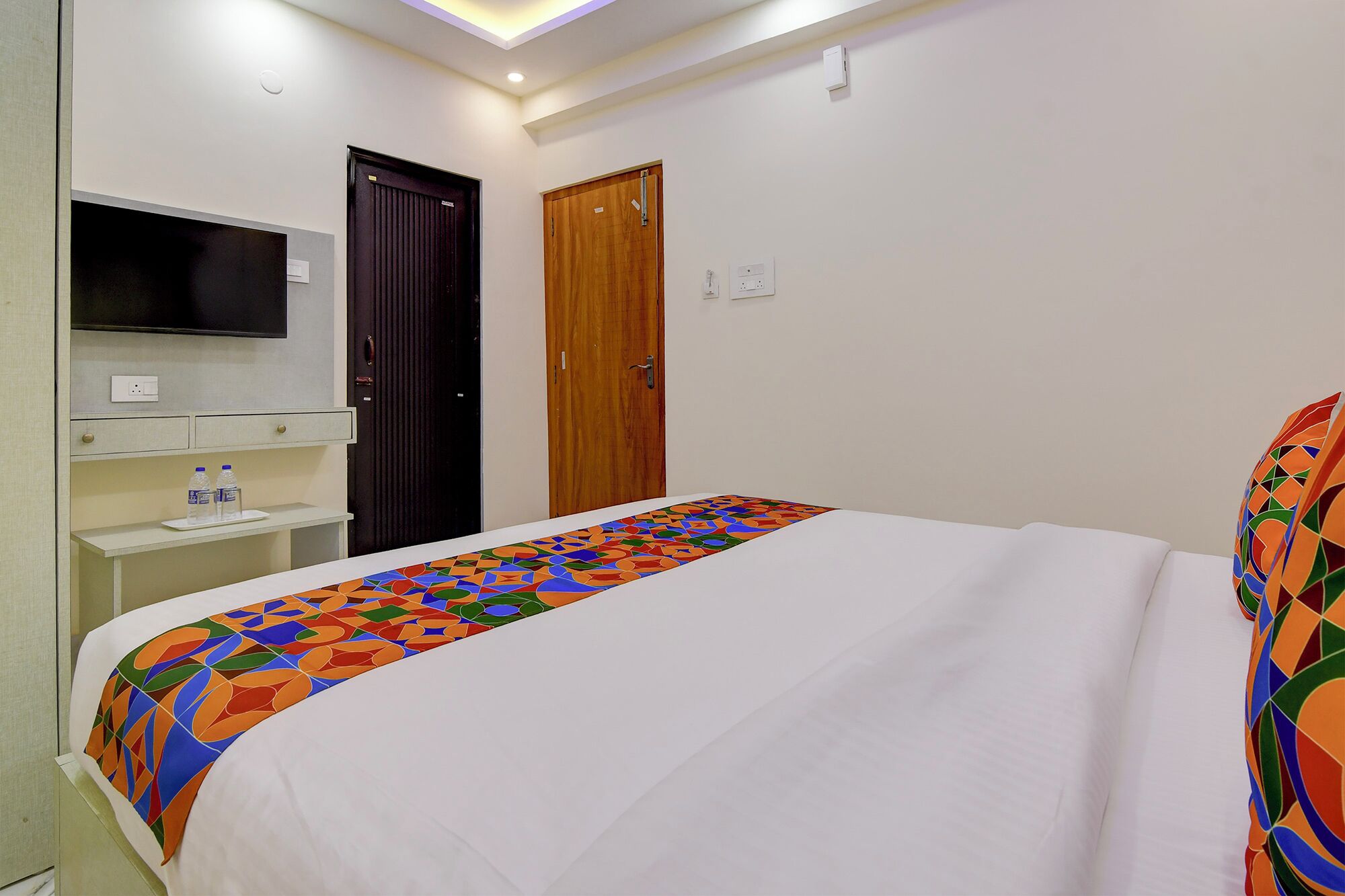 Una habitación de hotel con cama, televisor montado en la pared, puerta de madera y una pequeña mesa con una botella de agua.