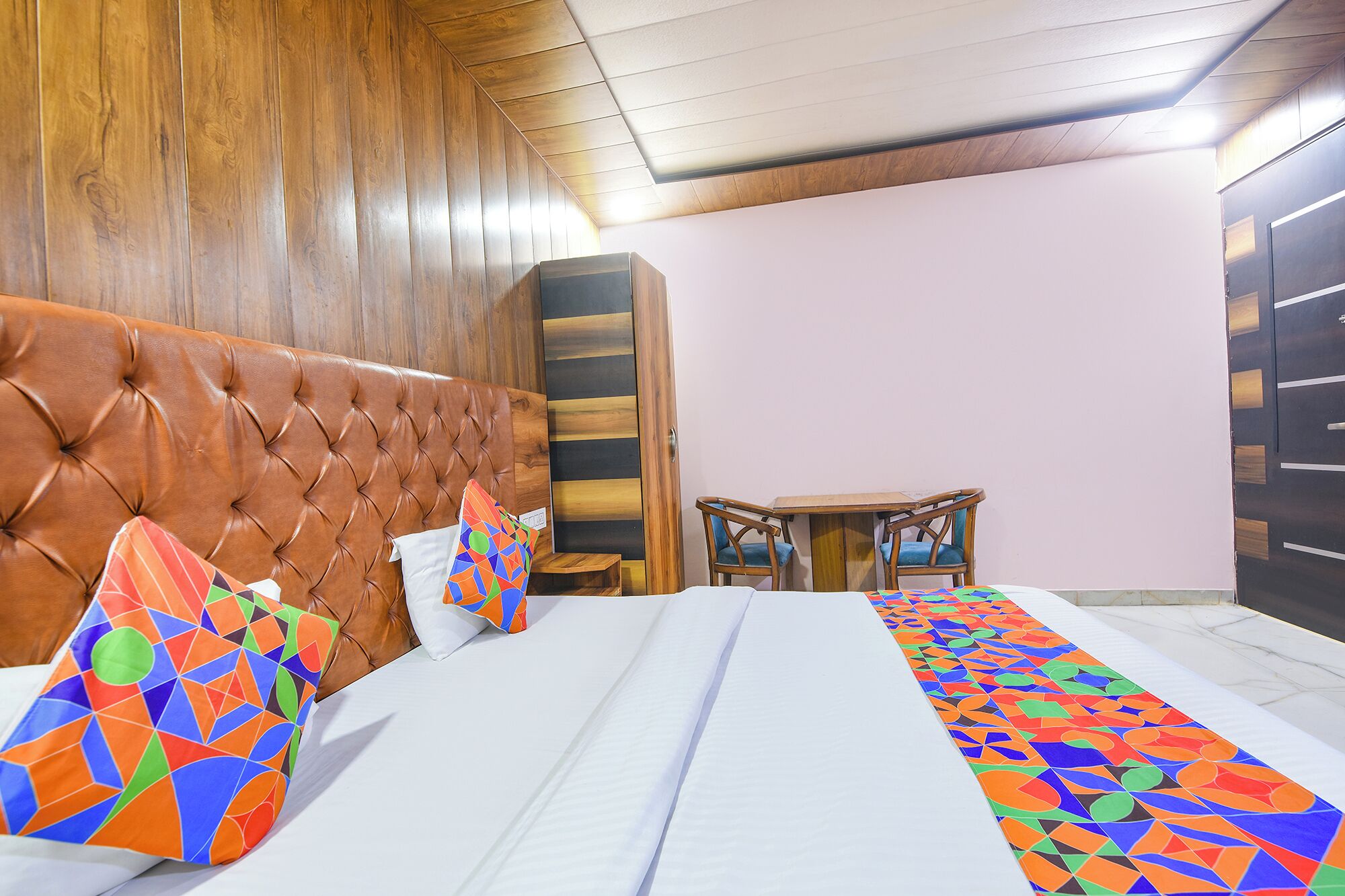 Une chambre d’hôtel avec une tête de lit capitonnée, un lit avec une literie à motifs, une petite table en bois, deux chaises et une armoire.