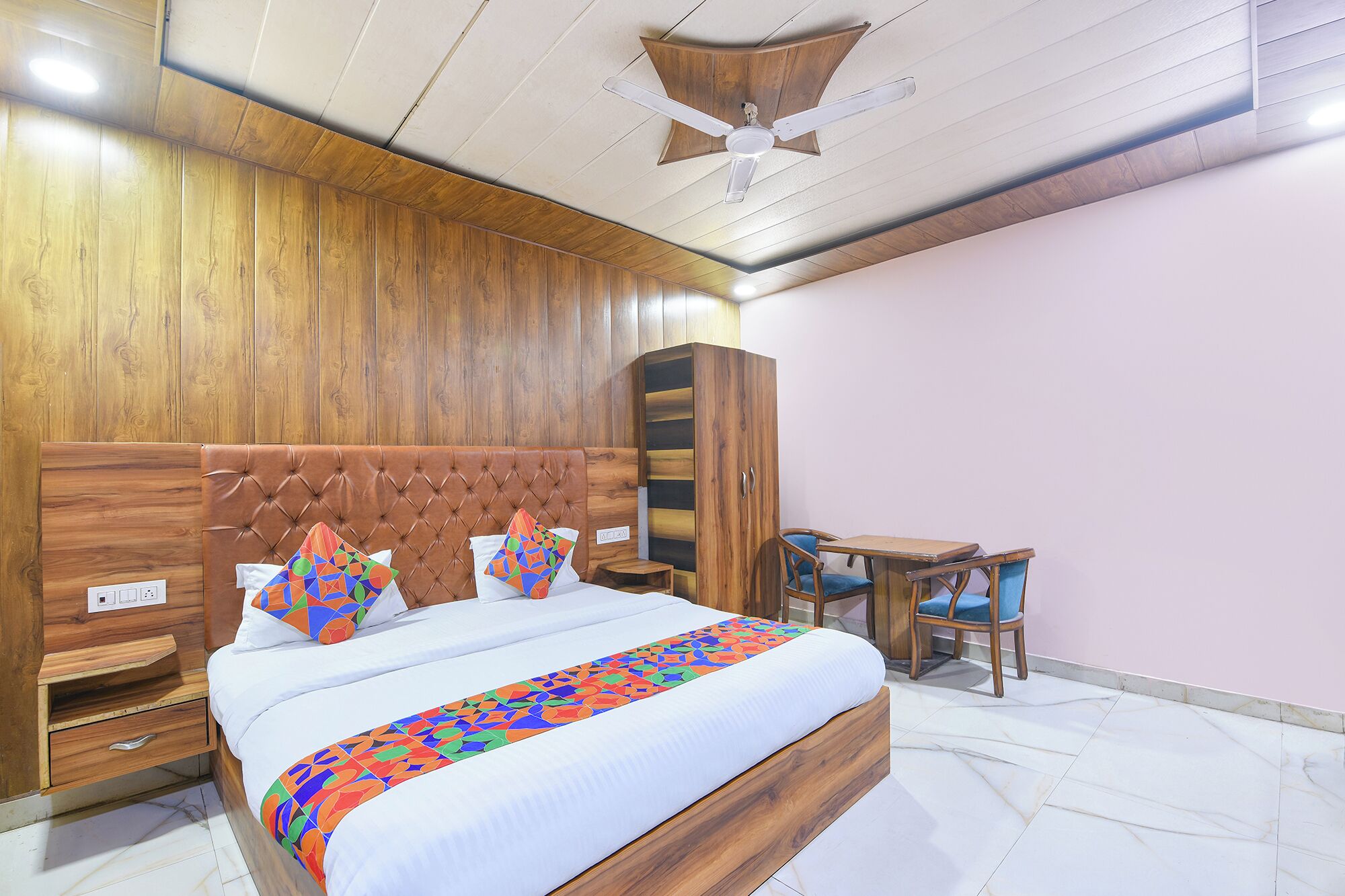 A bedroom with a wooden headboard, a bed with a colorful geometric bedspread, a nightstand, a wardrobe, a desk with a chair, and a ceiling fan.