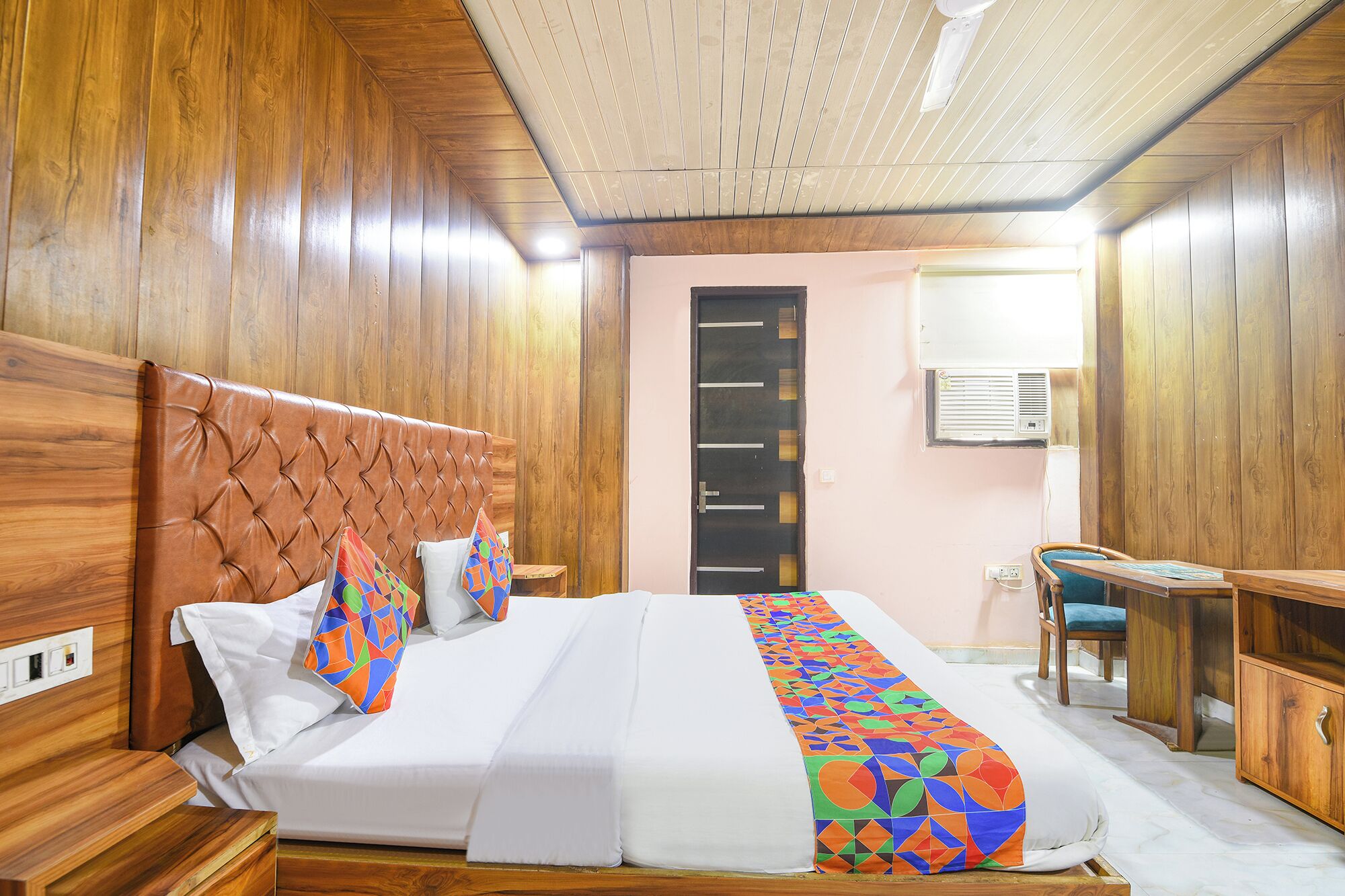 A hotel room with a wooden headboard, a bed with white linens, a colorful throw, a desk with a chair, and an air conditioning unit.
