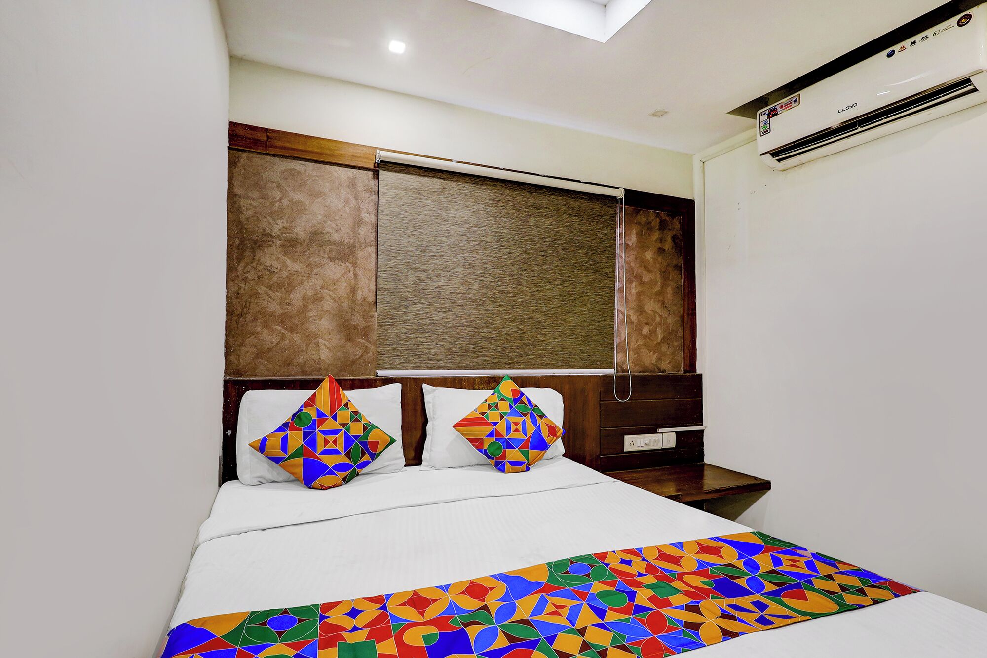 A hotel room with a bed, two pillows, a colorful bedspread, a wooden headboard, and an air conditioning unit.