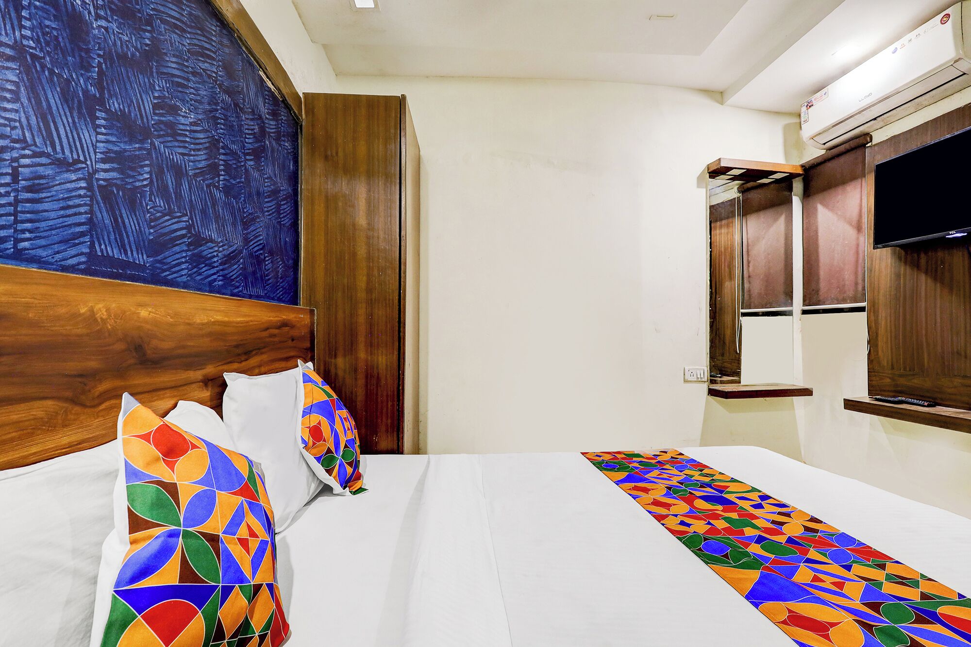 A hotel room with a bed, a wooden headboard, a large abstract painting, a television, and a window with curtains.