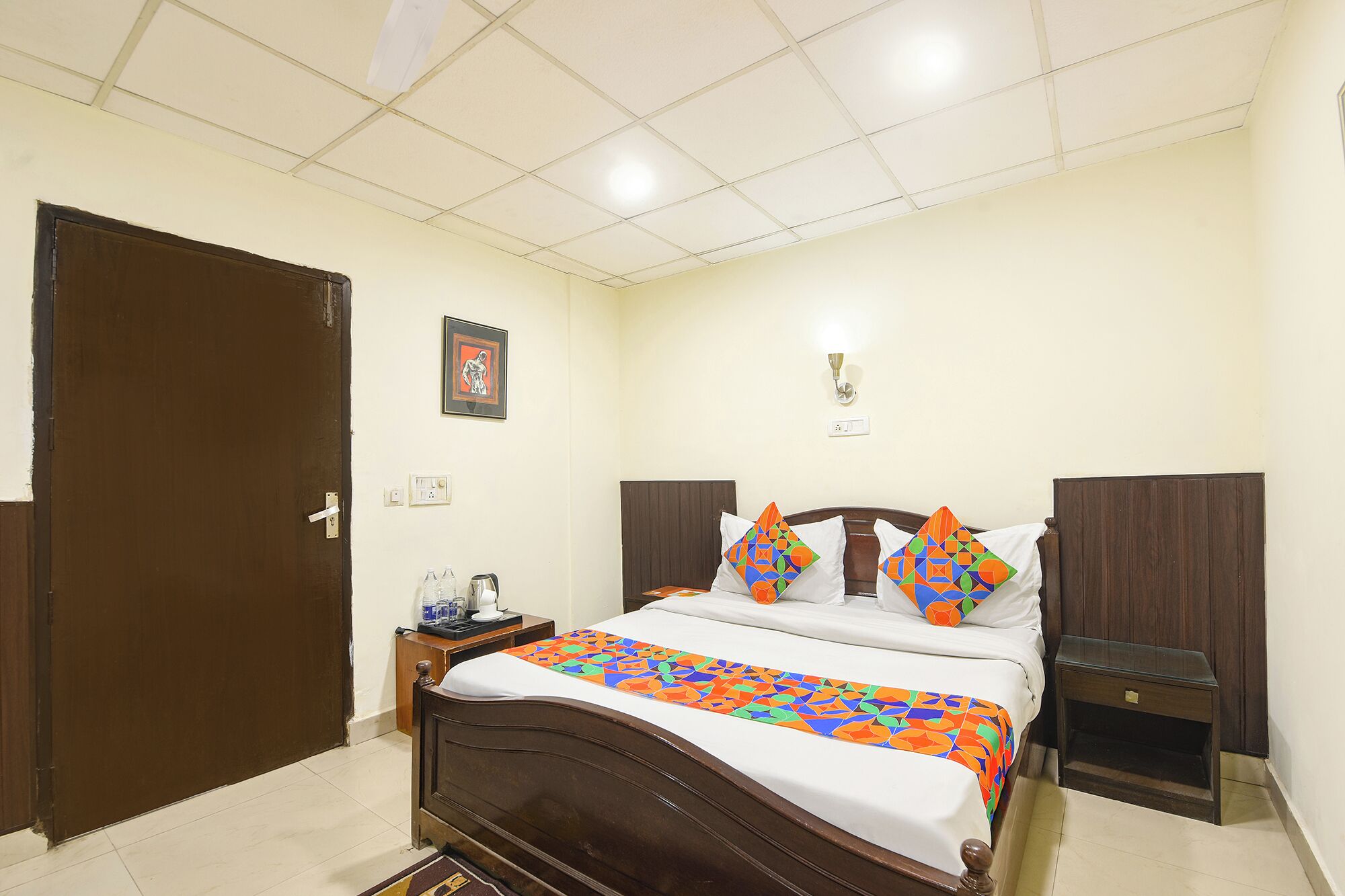 A bedroom with a wooden bed, a nightstand, a bedside table, a wall-mounted light, and a door.