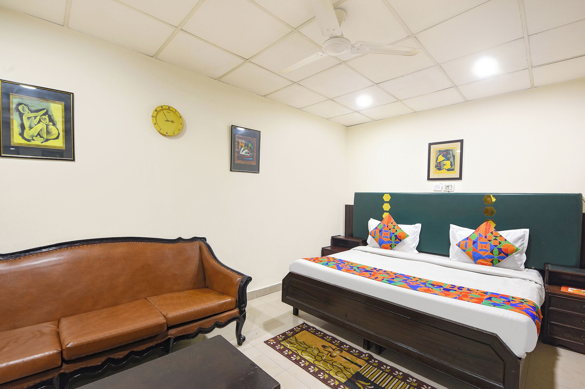 A bedroom with a bed, a brown leather sofa, a wooden nightstand, a wall clock, and framed artwork.