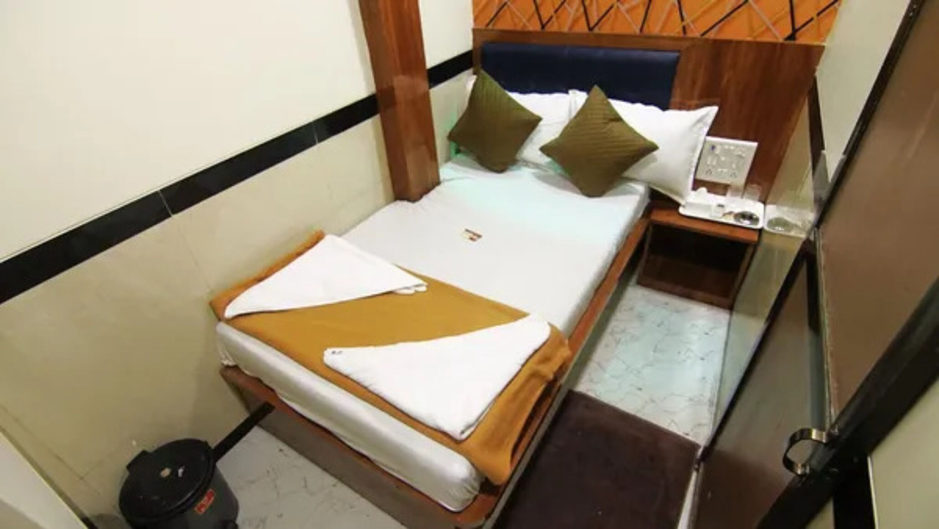 Quarto pequeno e compacto com cama, mesa de cabeceira e aquecedor fixado na parede.