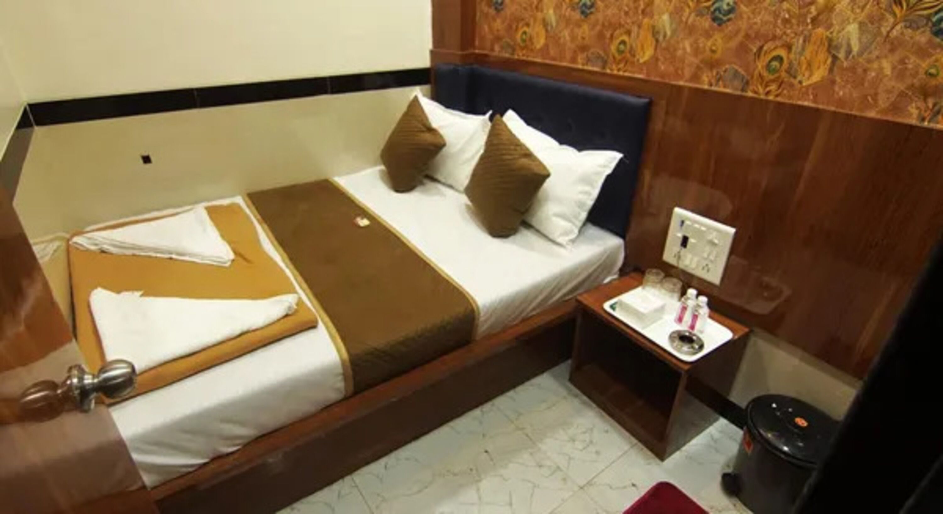 A hotel room with a bed, bedside table, and a trash bin.