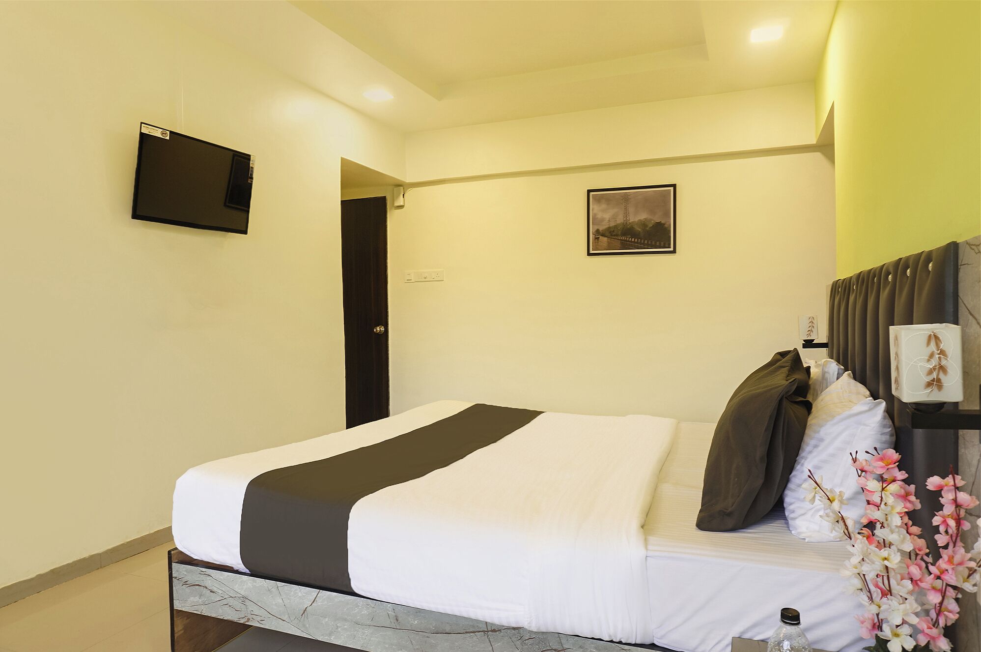A hotel room with a bed, a flat-screen TV, a framed picture on the wall, and a decorative plant.