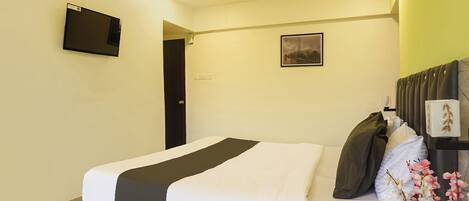 A hotel room with a bed, a flat-screen TV, a framed picture on the wall, and a decorative plant.