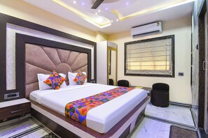 A modern hotel room with a large bed, a headboard, an air conditioning unit, a window with blinds, and a trash bin. - Fabhotel The Ruby Park (Kolkata)