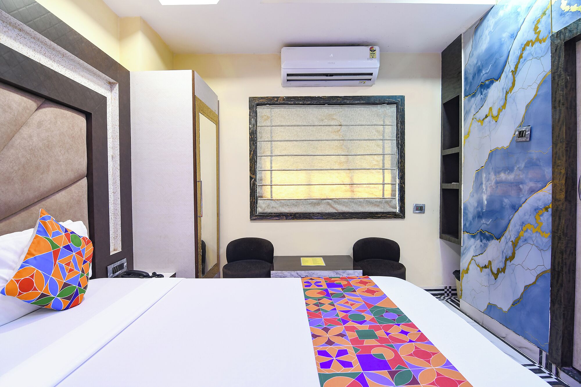 A hotel room with a bed, two chairs, a desk, an air conditioner, a window with blinds, and a large abstract painting on the wall.