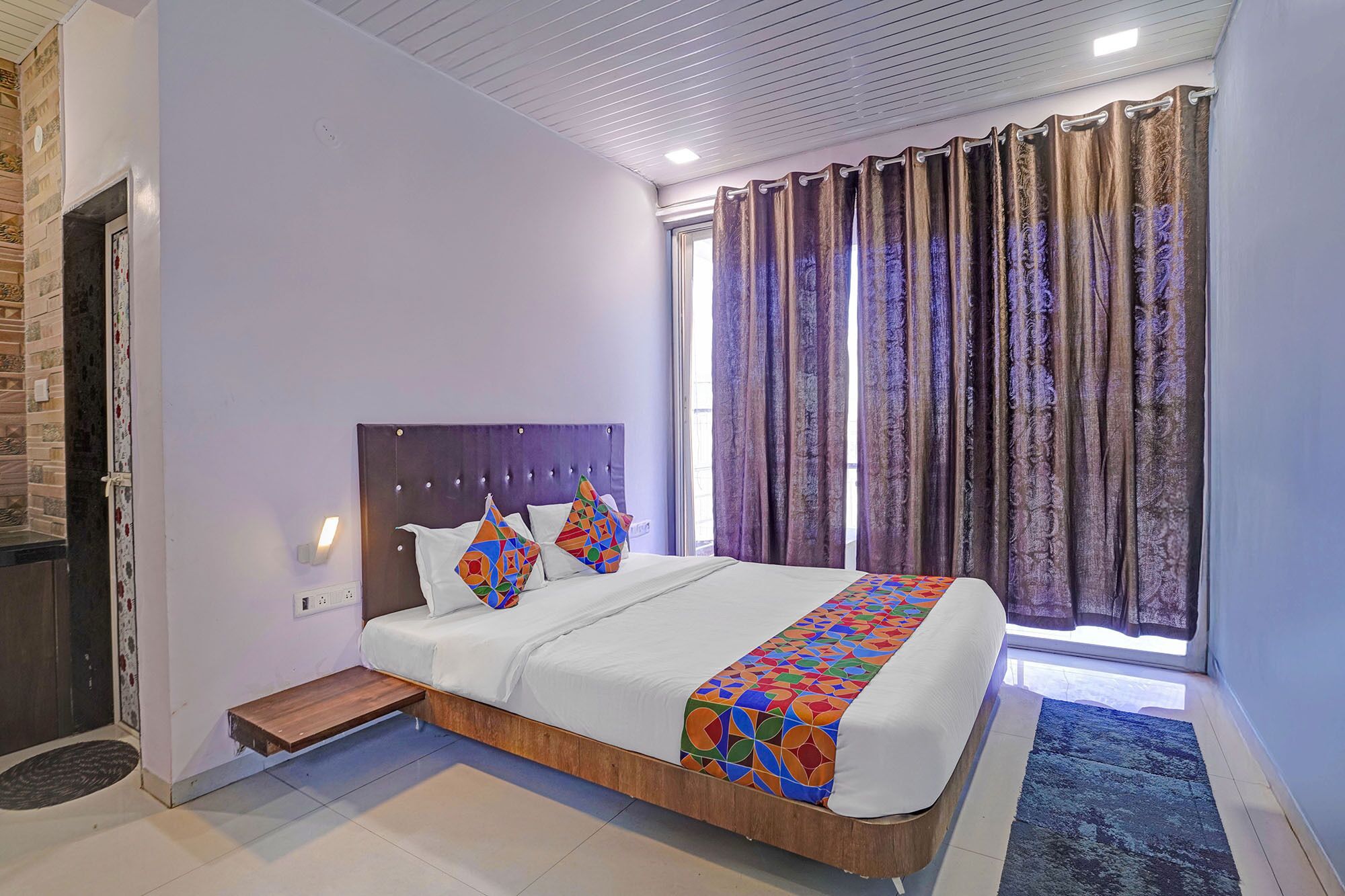 A bedroom with a bed, a headboard, a small wooden bench, and a window with curtains.