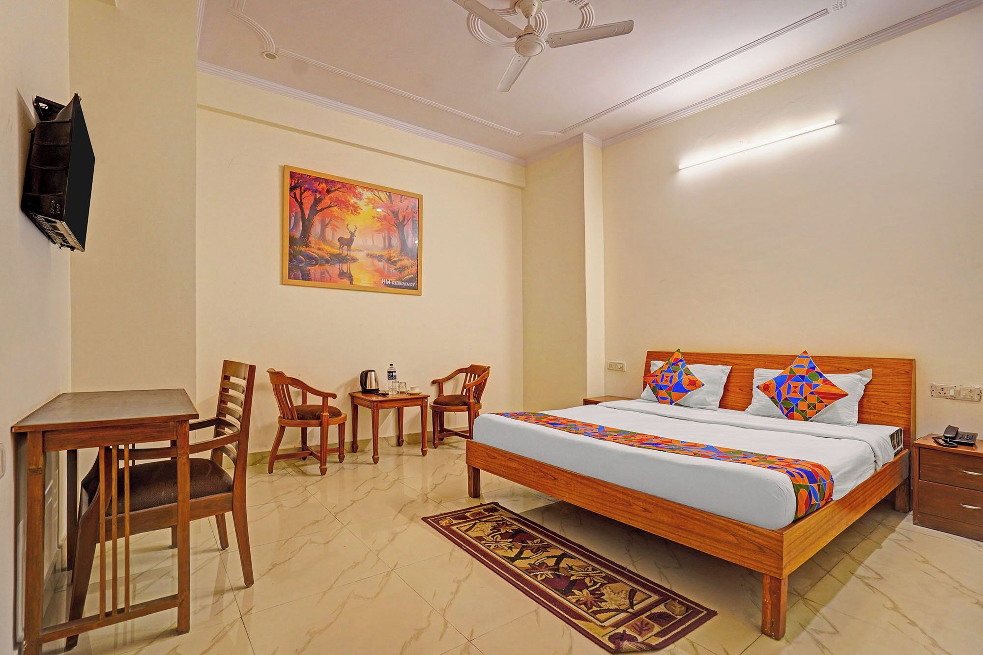 A bedroom with a bed, a desk, two chairs, a television, and a painting on the wall.