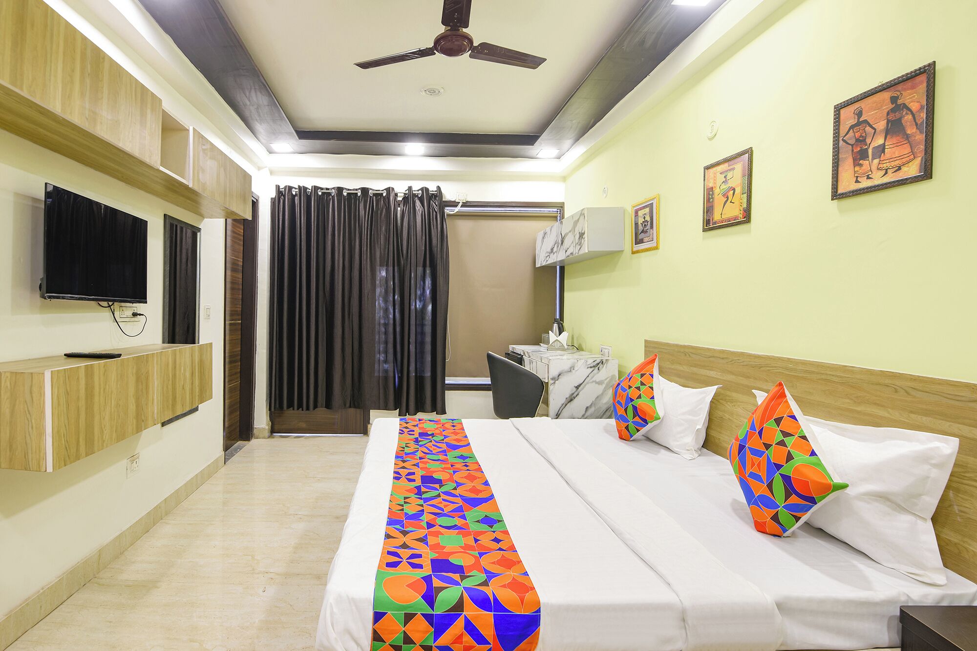 A hotel room with a bed, a television, a ceiling fan, and artwork on the walls.