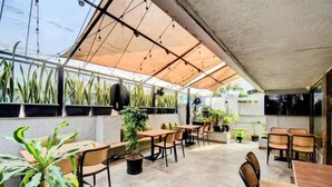 Un patio al aire libre con mesas y sillas, plantas en macetas y una estructura de techo de cristal.