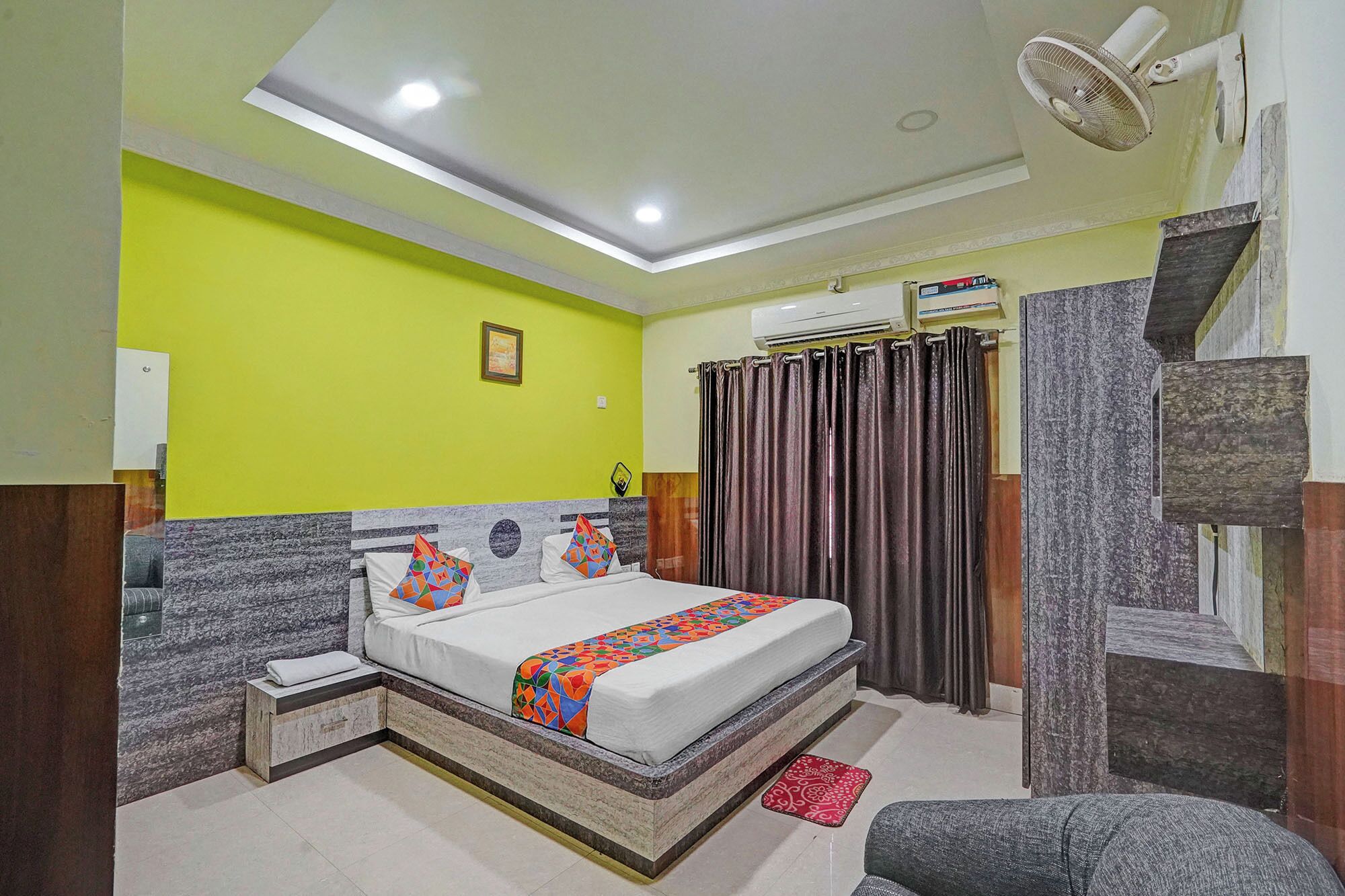 Une chambre à coucher comprenant un lit, une table de chevet, un miroir, un ventilateur de plafond et un appareil de climatisation.
