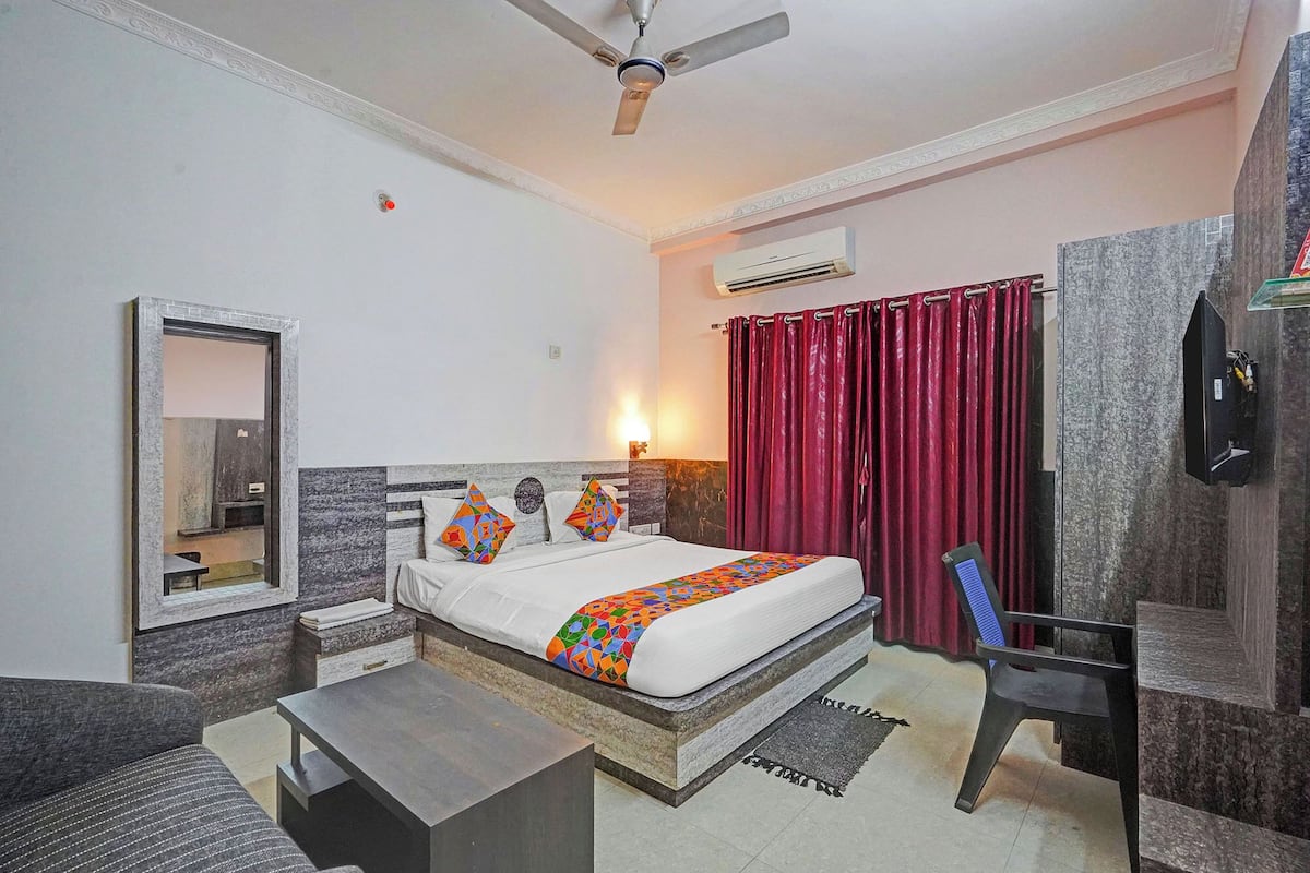A hotel room with a bed, a desk, a chair, a ceiling fan, an air conditioning unit, a mirror, and a red curtain.