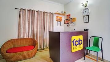 A reception desk with a sign for 'Tab Hotels,' a wicker chair with a red cushion, a green chair, and a clock on the wall.