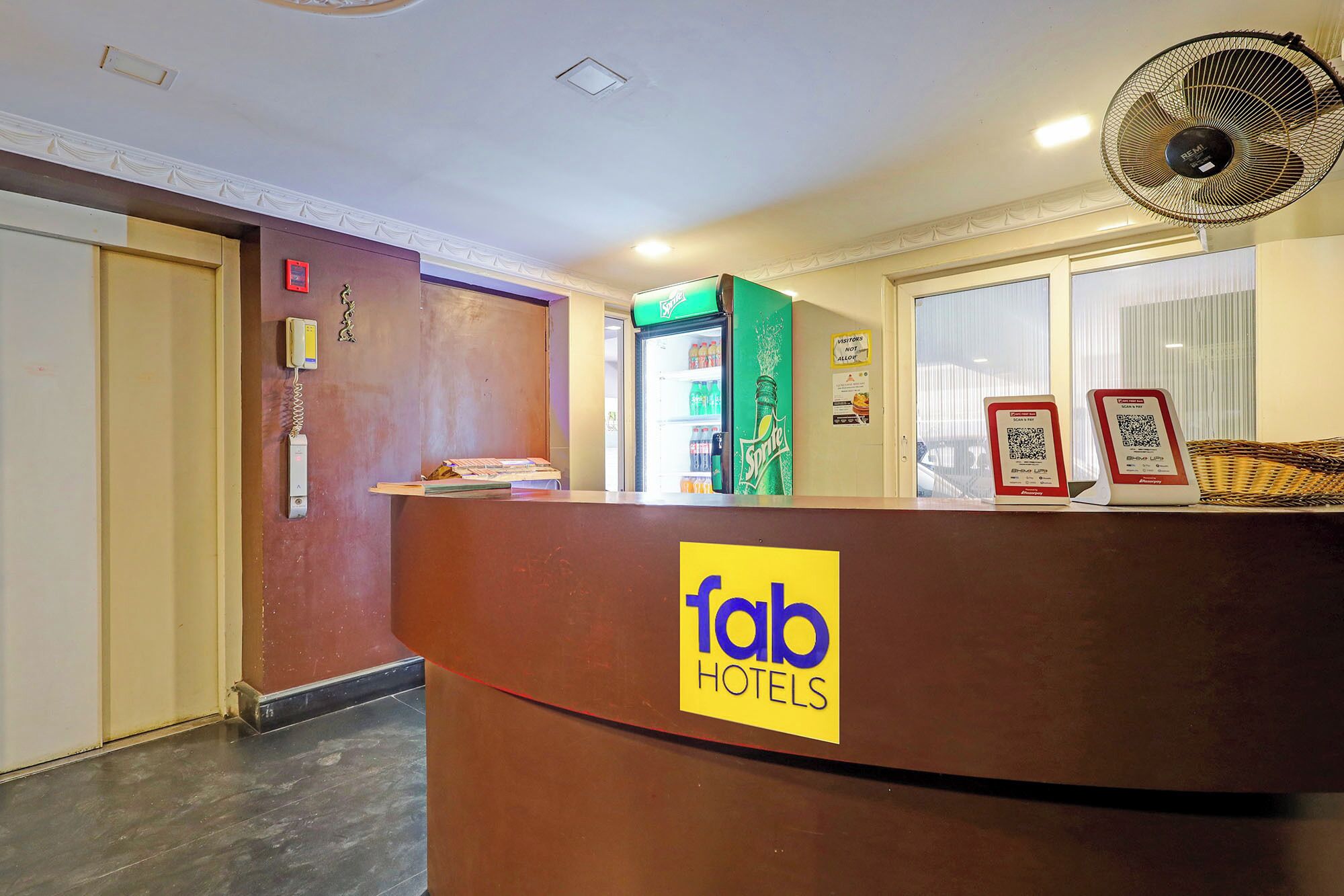 A hotel reception desk with a sign for 'Tab Hotels,' a refrigerator stocked with beverages, a fan, and a QR code for check-in.
