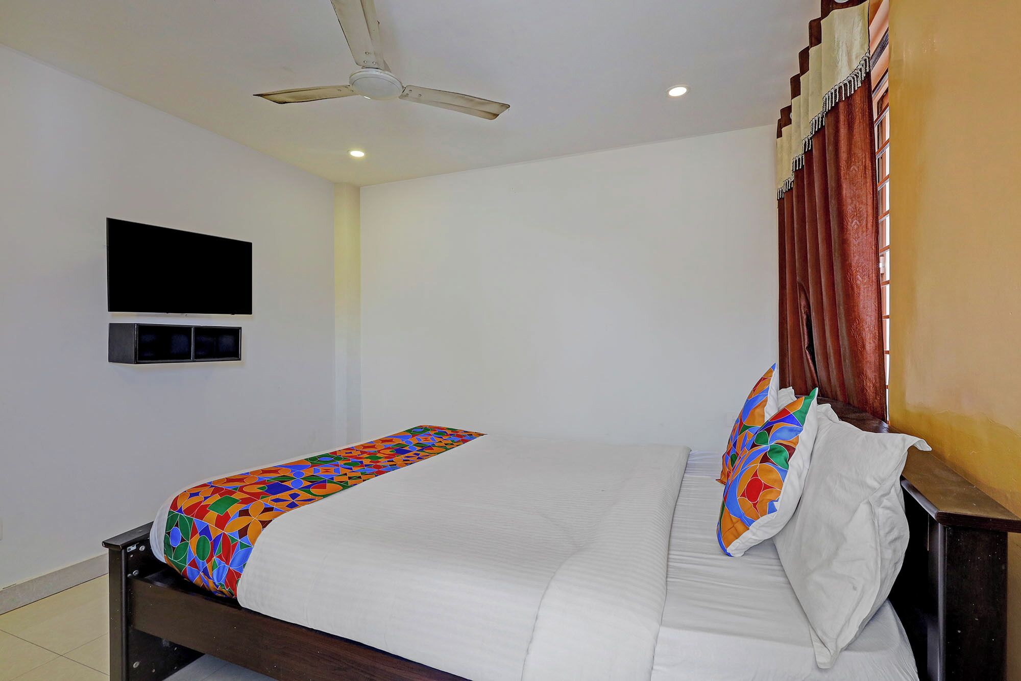 A bedroom with a bed, a ceiling fan, a TV mounted on the wall, and a window with curtains.
