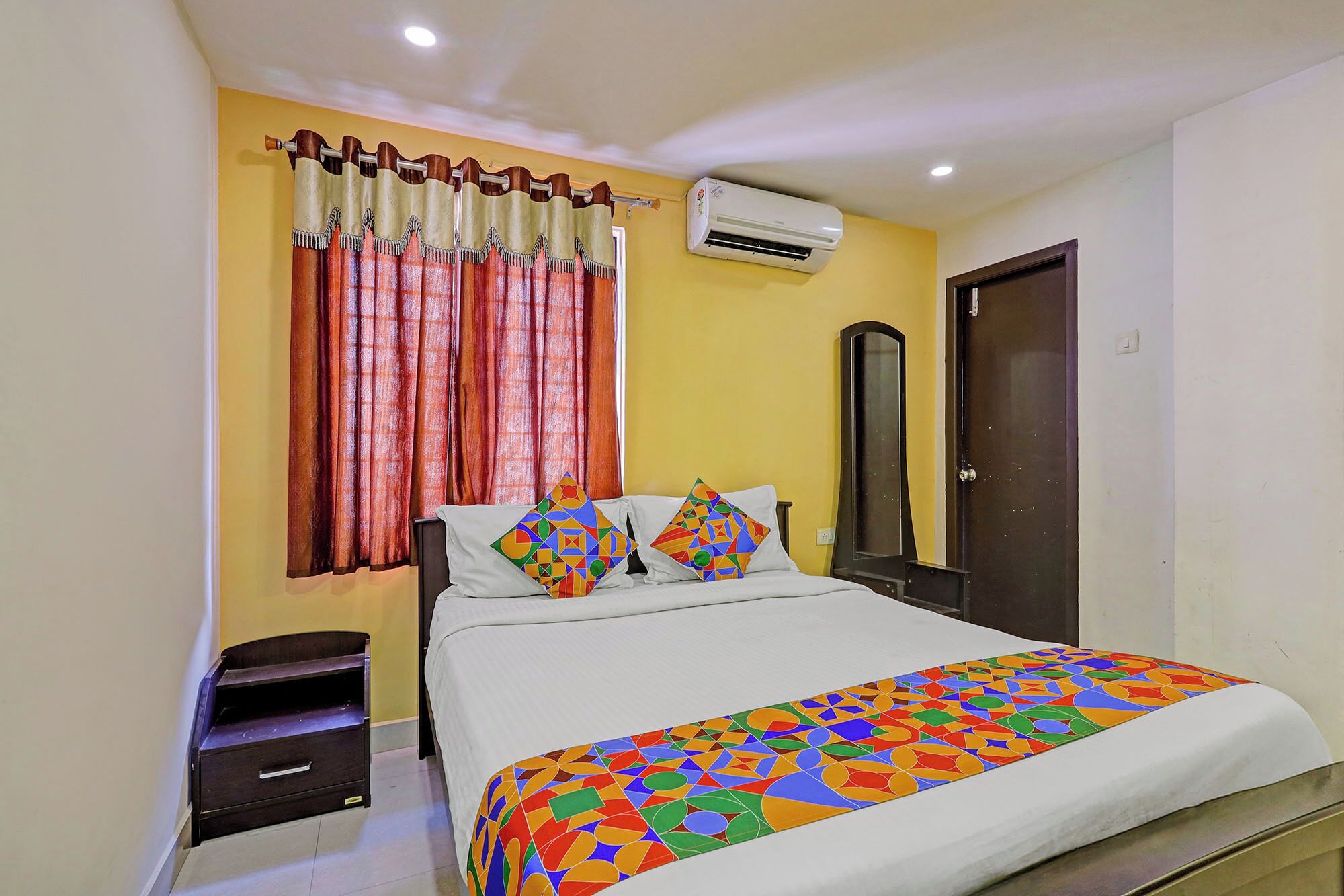 A bedroom with a bed, a nightstand, a wardrobe, a mirror, and an air conditioning unit.