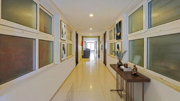 A hallway with large windows, a polished floor, and a desk with decorative items. - Fabhotel Neha (Chennai)