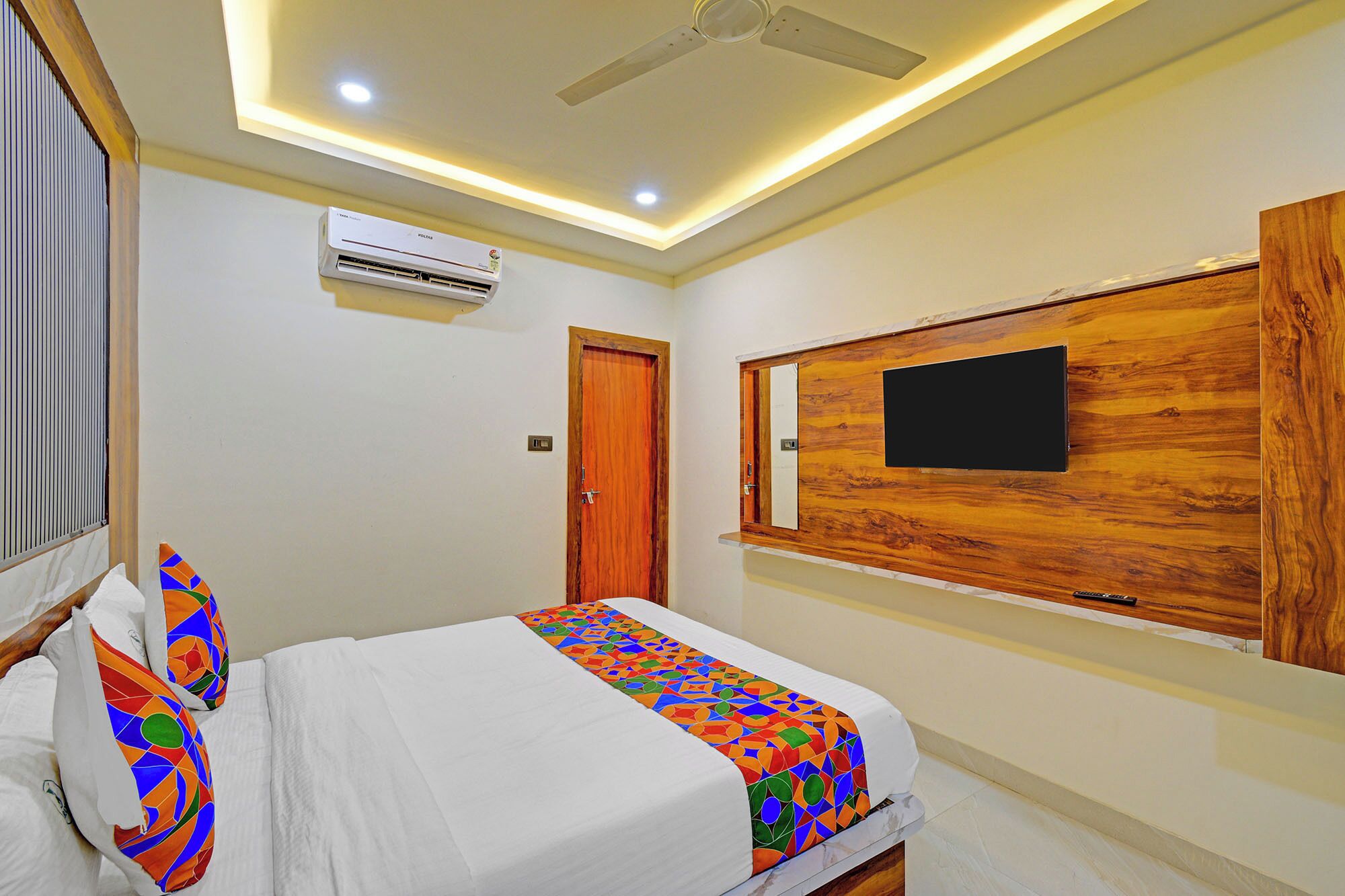 Un dormitorio con cama, ventilador de techo, unidad de aire acondicionado, cabecera de madera con televisor y una puerta.