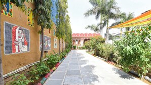 A pathway lined with colorful murals, palm trees, and a building with a red roof in the background. - Fabhotel Nimabal Dhani (Jaipur)