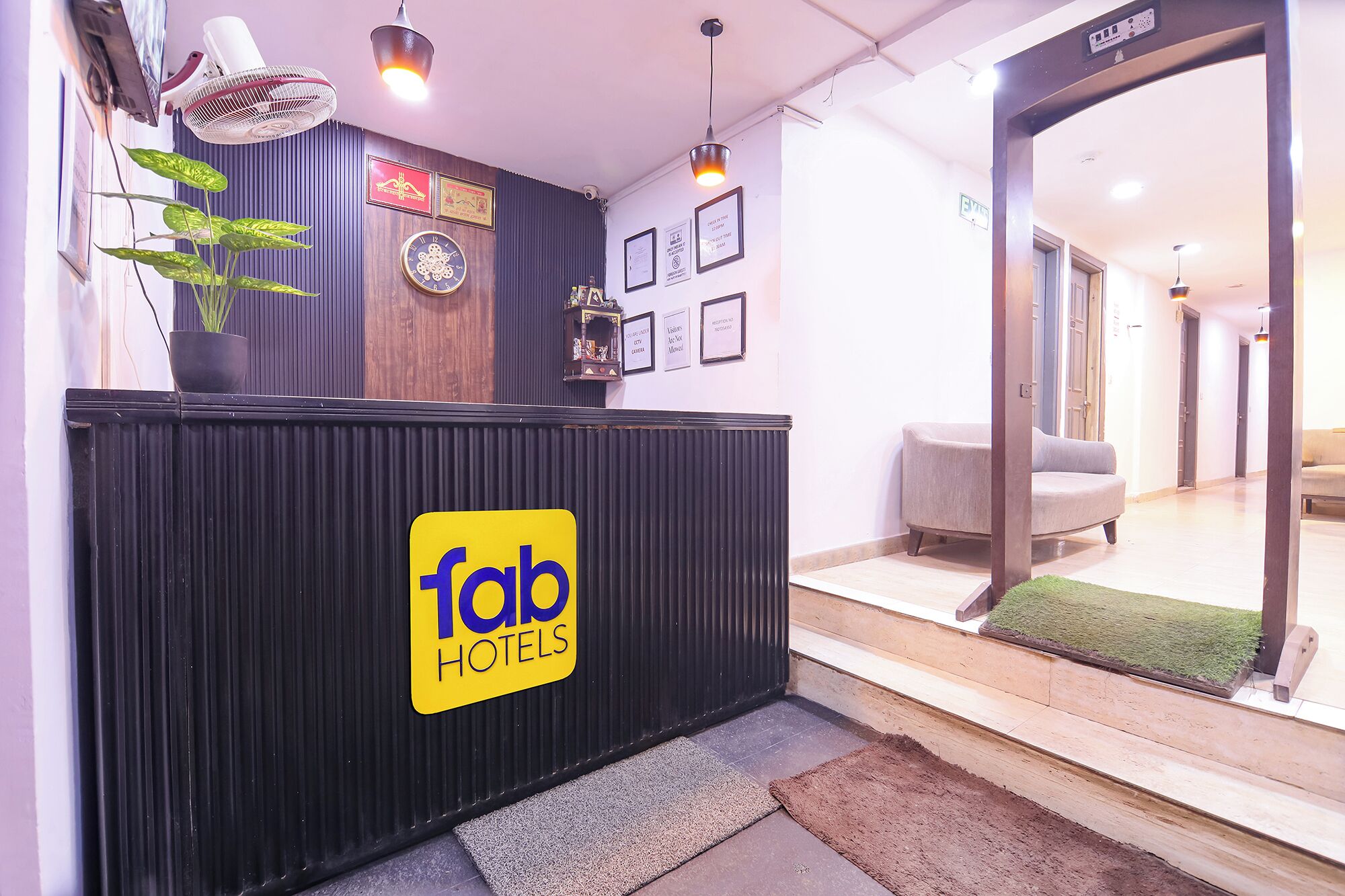 A reception desk with a sign for 'fab hotels,' a clock, framed certificates, and a plant.
