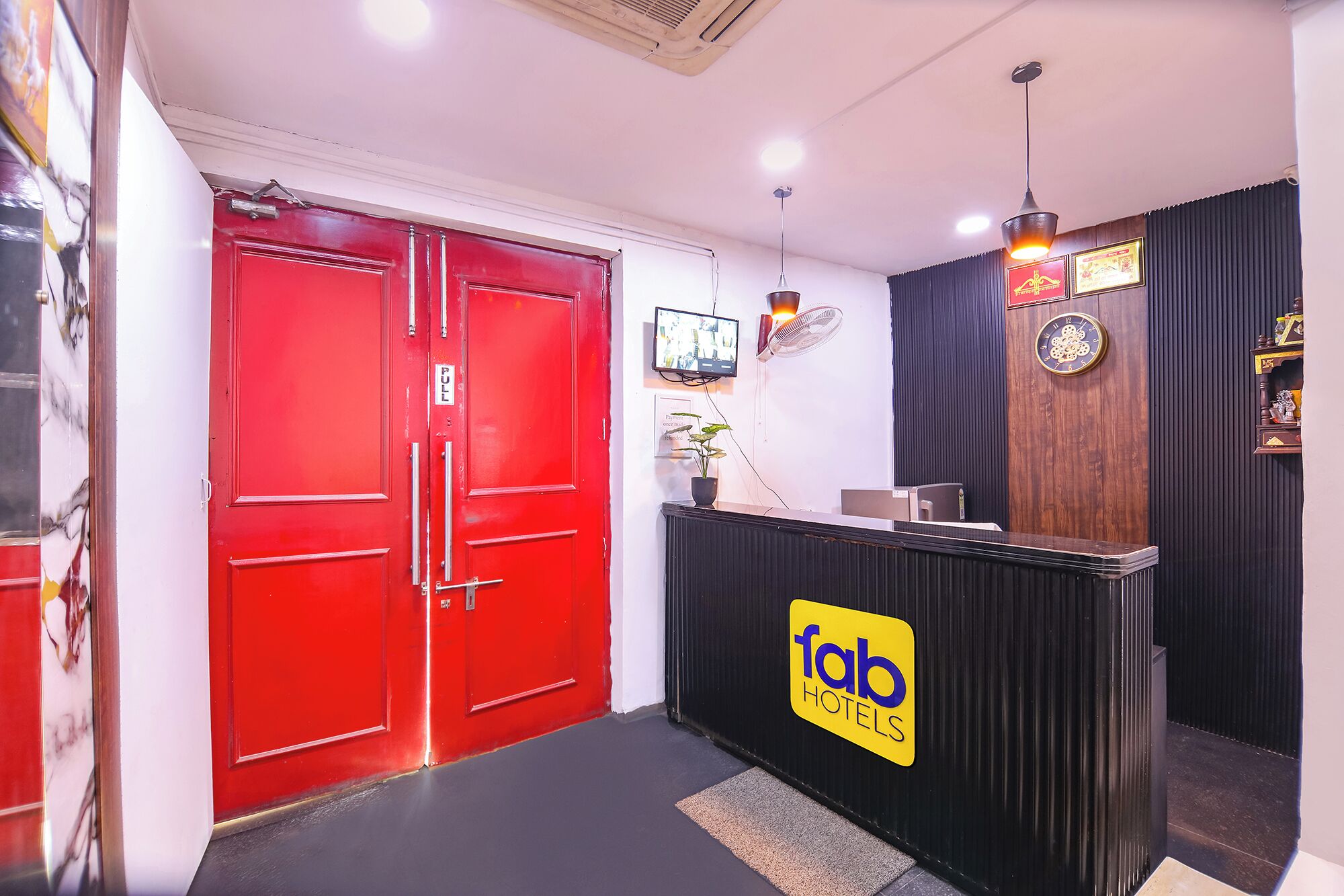 A hotel reception area with a red door, a reception desk with a 'fab hotels' sign, a clock, and a wall-mounted fan.