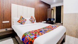 Quarto de hotel com cabeceira de madeira, cama com colcha de padrão geométrico colorido, mesa de cabeceira com abajur e criado-mudo com controle remoto.