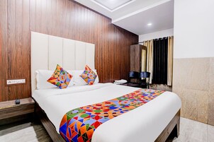 A hotel room with a wooden headboard, a bed with a colorful geometric-patterned bedspread, a bedside table with a lamp, and a nightstand with a remote control.