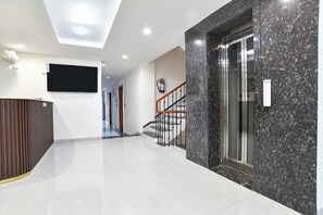 A modern hotel lobby with a marble reception desk, a staircase with a glass elevator, and a fire extinguisher mounted on the wall. - Fabhotel The Backyard Bowl (Sanwer)