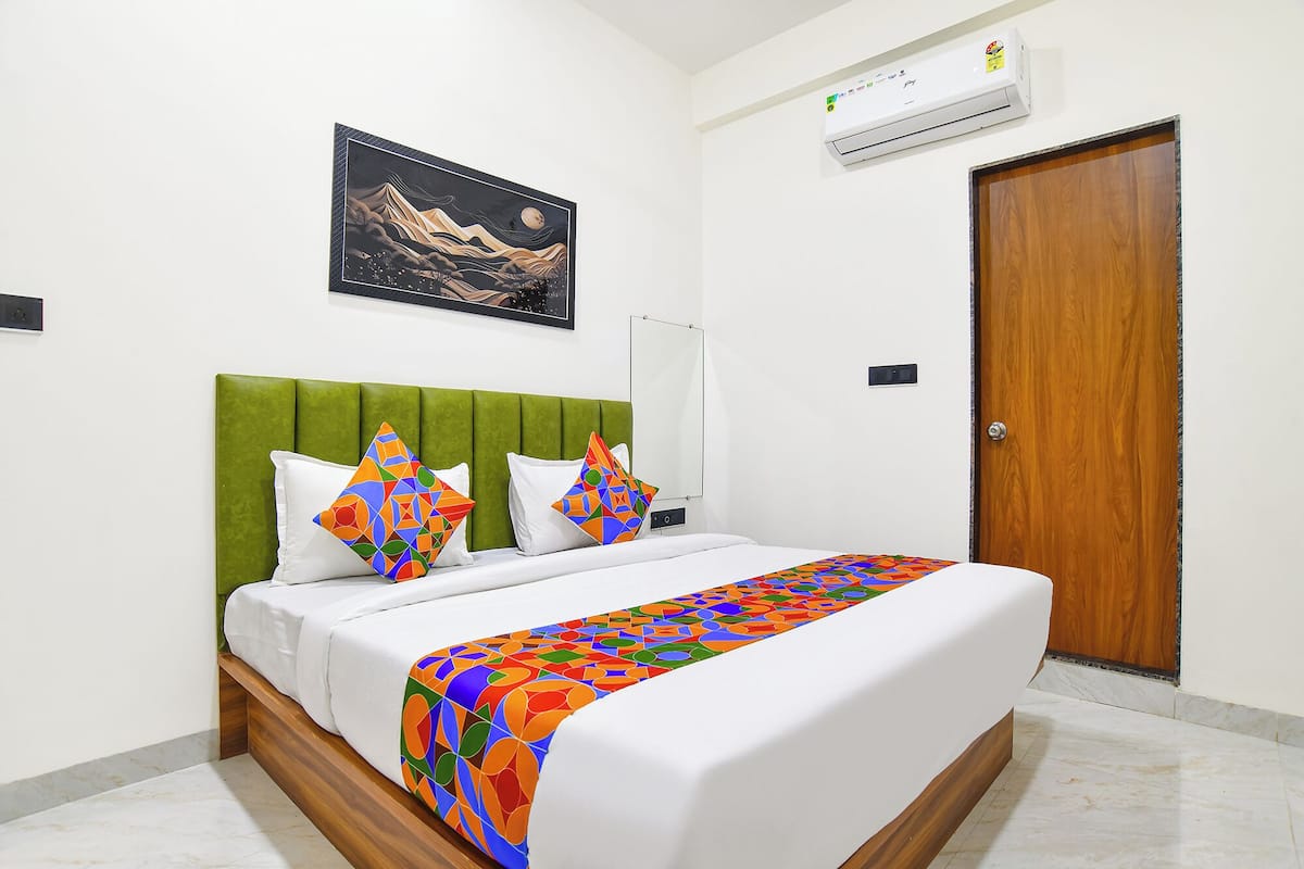 Una habitación de hotel ordenada con una cama, cabecera tapizada de verde, aire acondicionado montado en la pared y una puerta de madera.