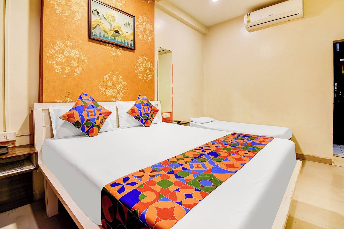 A hotel room with a bed, a nightstand, a mirror, an air conditioner, and a painting on the wall.