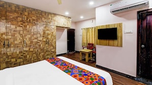 Une chambre d’hôtel avec une armoire en bois, un lit recouvert d’une courtepointe colorée, une petite table, une chaise, un téléviseur fixé au mur et un appareil de climatisation.