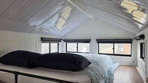 Une chambre avec un plafond en pente, deux fenêtres équipées de stores, un lit avec une literie sombre et un radiateur.