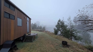 Une cabane en bois avec une terrasse, un banc et un foyer, située au bord d’un lac.