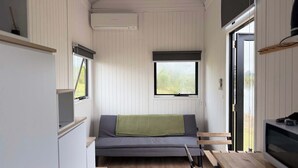 Et kompakt værelse med sofa, et lille bord, et fjernsyn og et vindue med udsigt til grøn bevoksning.