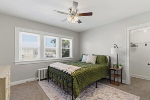 A bedroom with a bed, a radiator, a window with blinds, a ceiling fan, and a door.